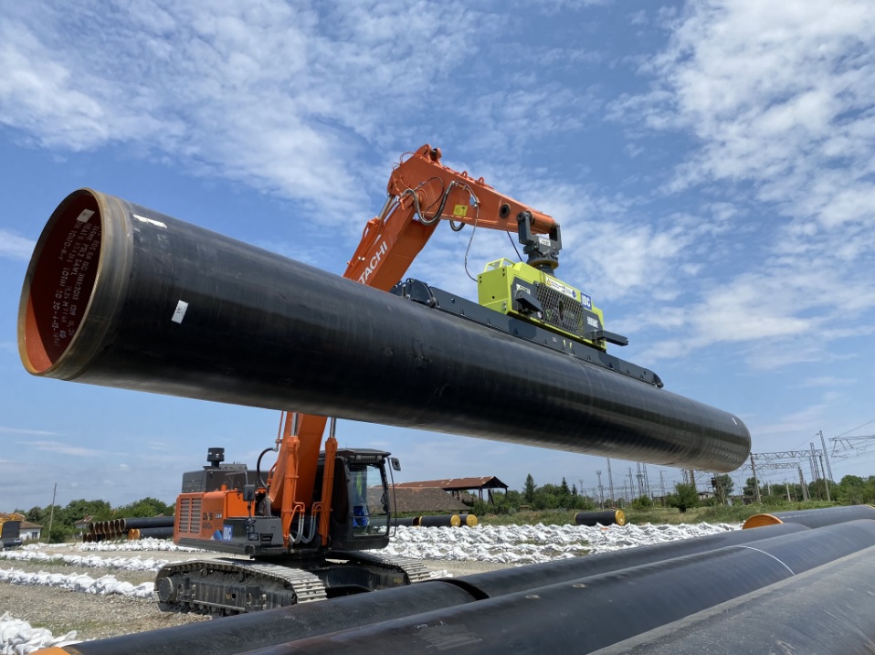 An orange Hitachi excavator utilizing a Vlentec vacuum lifter for efficient pipe lifting of large diameter coated pipes.