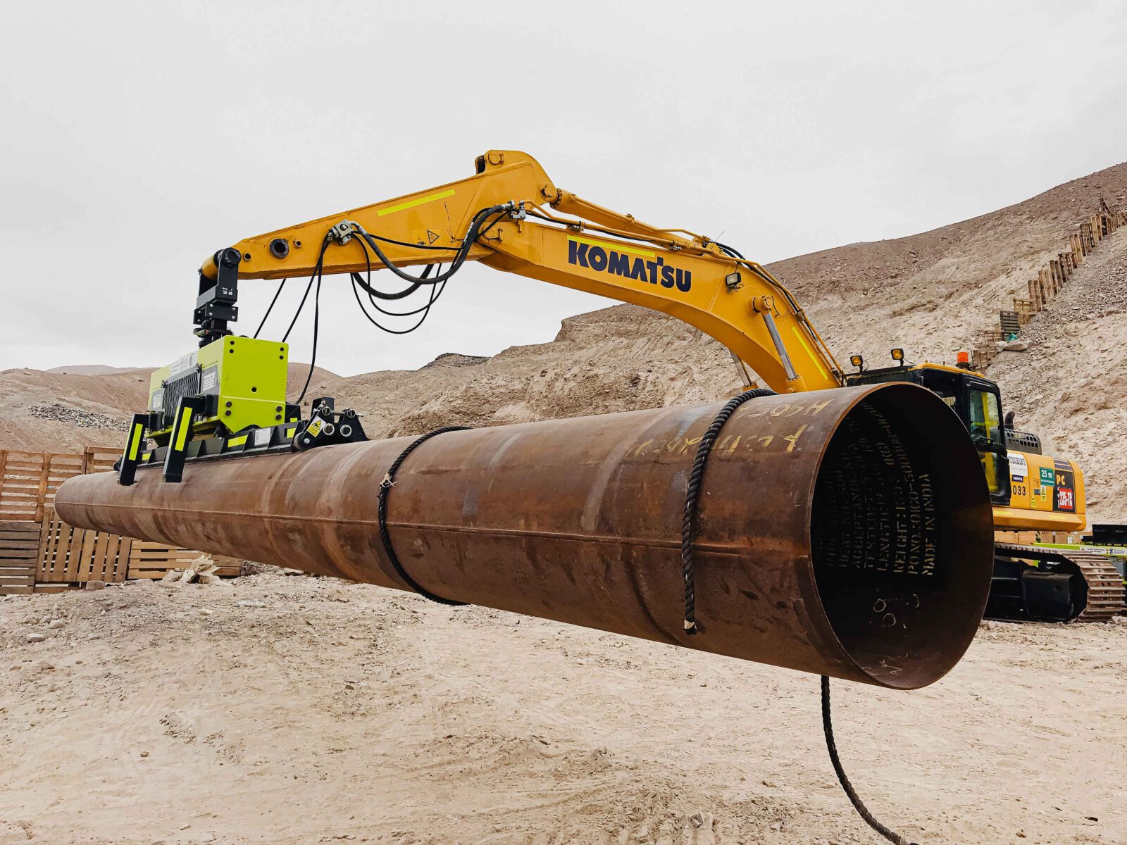 A yellow komatsu excavator using a vlentec vacuum lifter for heavy-duty pipe lifting of a large diameter steel pipe in a desert environment.