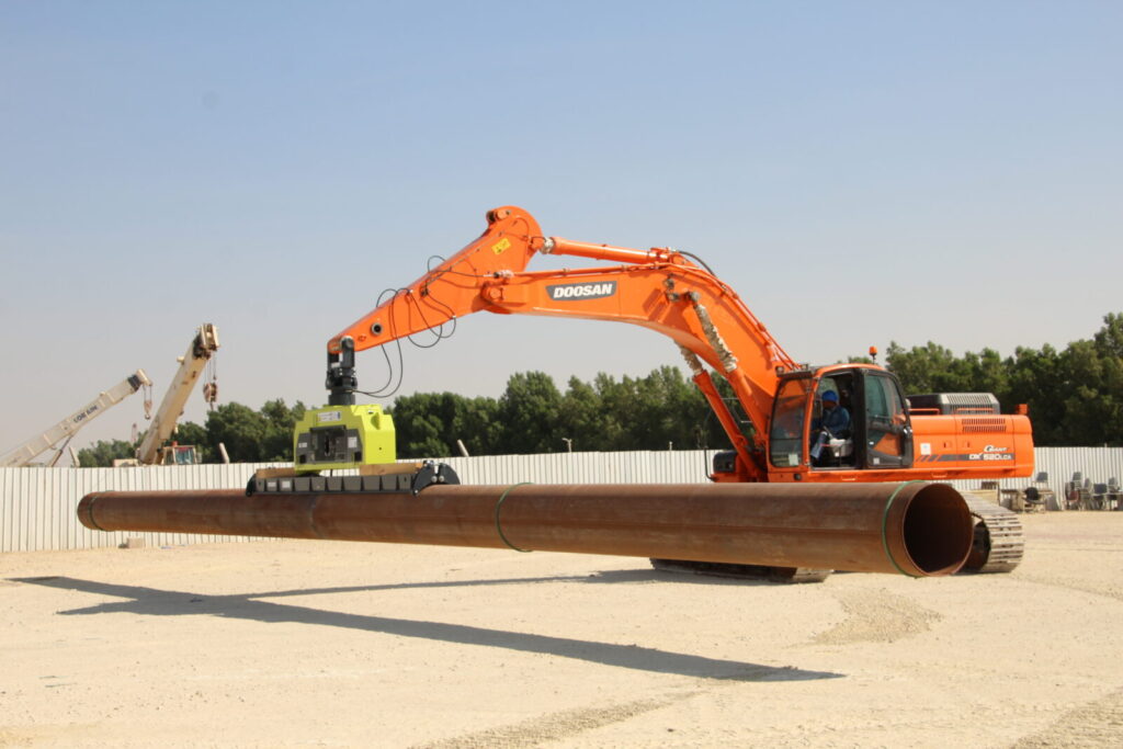 A doosan excavator using a vlentec vacuum lifter to handle a large diameter steel pipe on a construction site.