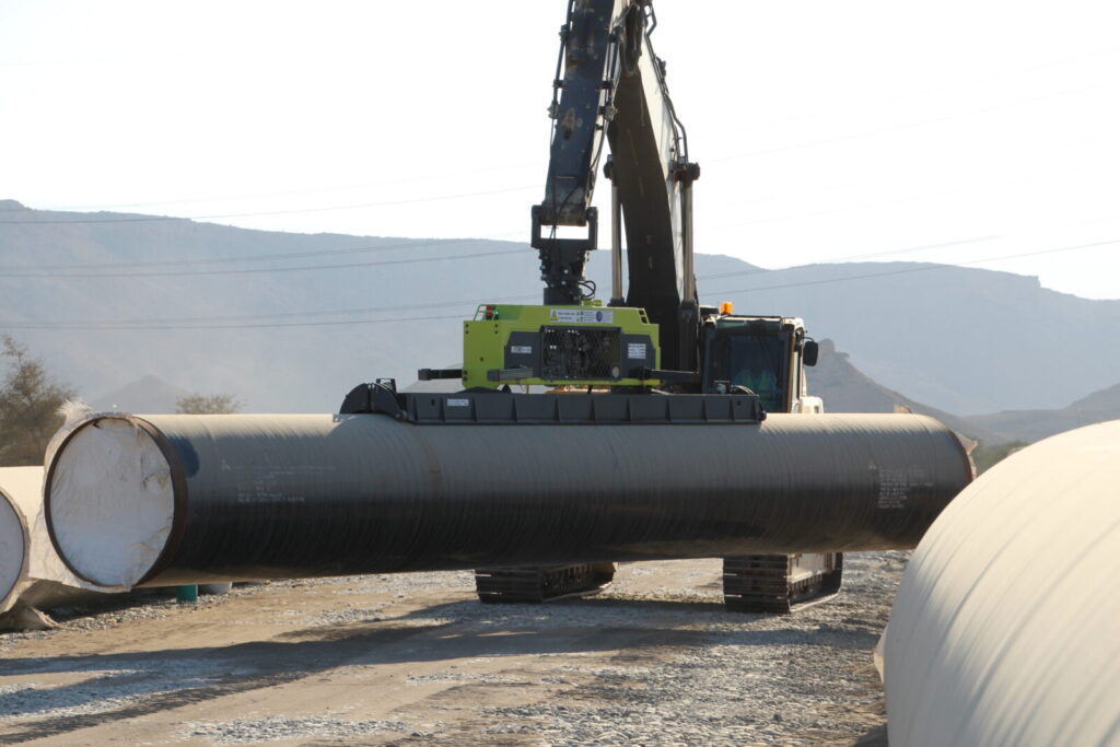 An excavator using a vlentec vacuum lifter for precise pipe lifting and handling of large diameter pipes in a rugged landscape.